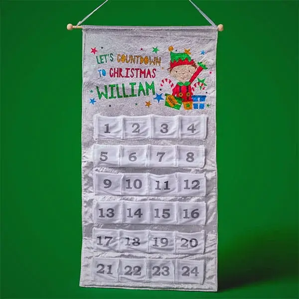 Personalised Christmas countdown calendar with numbered pockets, festive elf illustration, and custom name displayed on a hanging fabric wall banner.